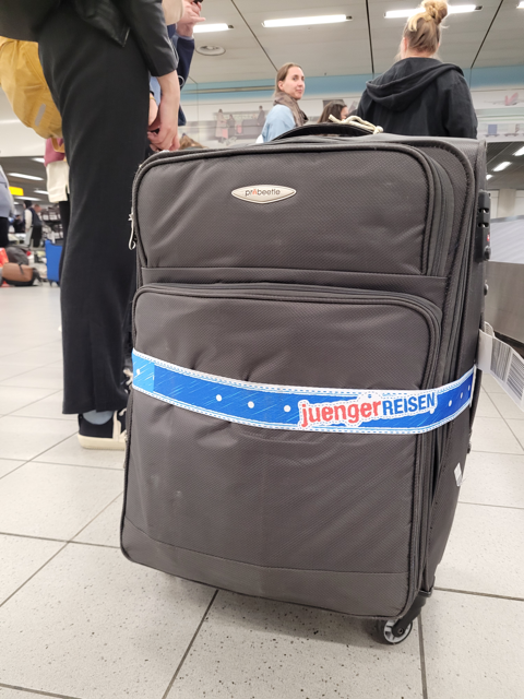 Großer Reisekoffer mit juengerREISEN-Koffergurt am Gepäckband, im Hintergrund wartende Menschen.