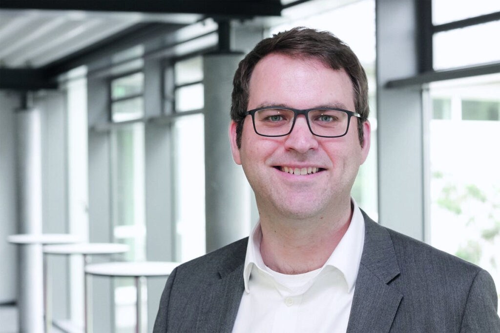 Porträt eines Mannes mit Brille und kurzem Haar in grauem Sakko und weißem Hemd vor hellem, modernem Hintergrund.