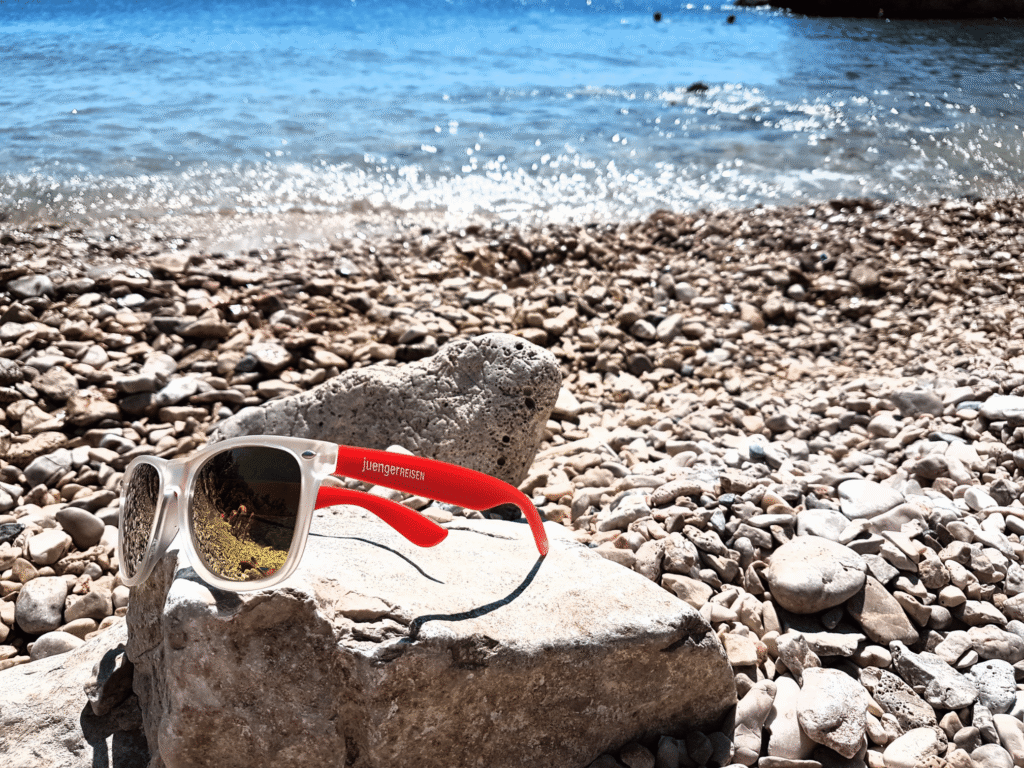 Sonnenbrille mit roten Bügeln liegt auf Felsen am steinigen Strand, dahinter glitzert das blaue Meer.