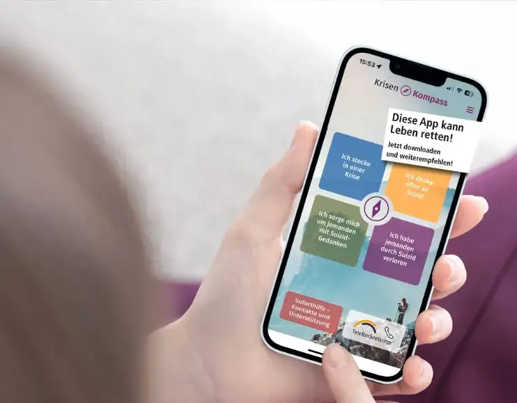 Smartphone in einer Hand zeigt die Startseite der App „Krisenkompass“ mit mehreren farbigen Auswahlfeldern für Unterstützung bei persönlichen Krisen und suizidalen Gedanken.