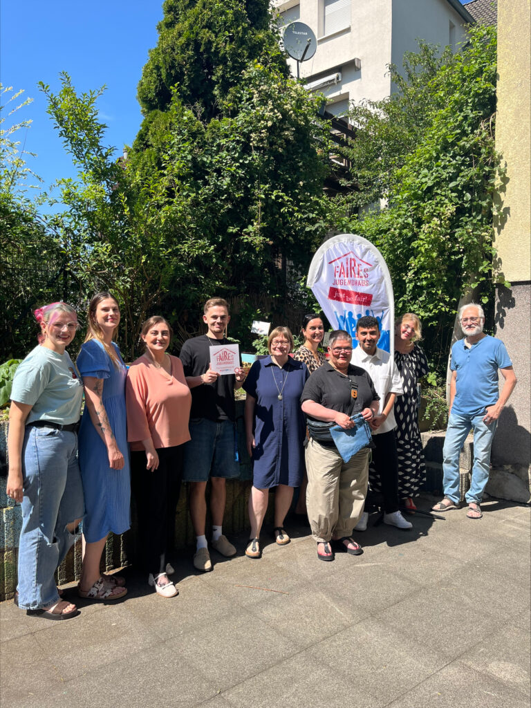 Gruppenfoto vor dem Café Frosch: Mehrere Personen stehen bei sonnigem Wetter vor dem Kinder- und Jugendzentrum und präsentieren die Auszeichnung als „Faires Jugendhaus“.