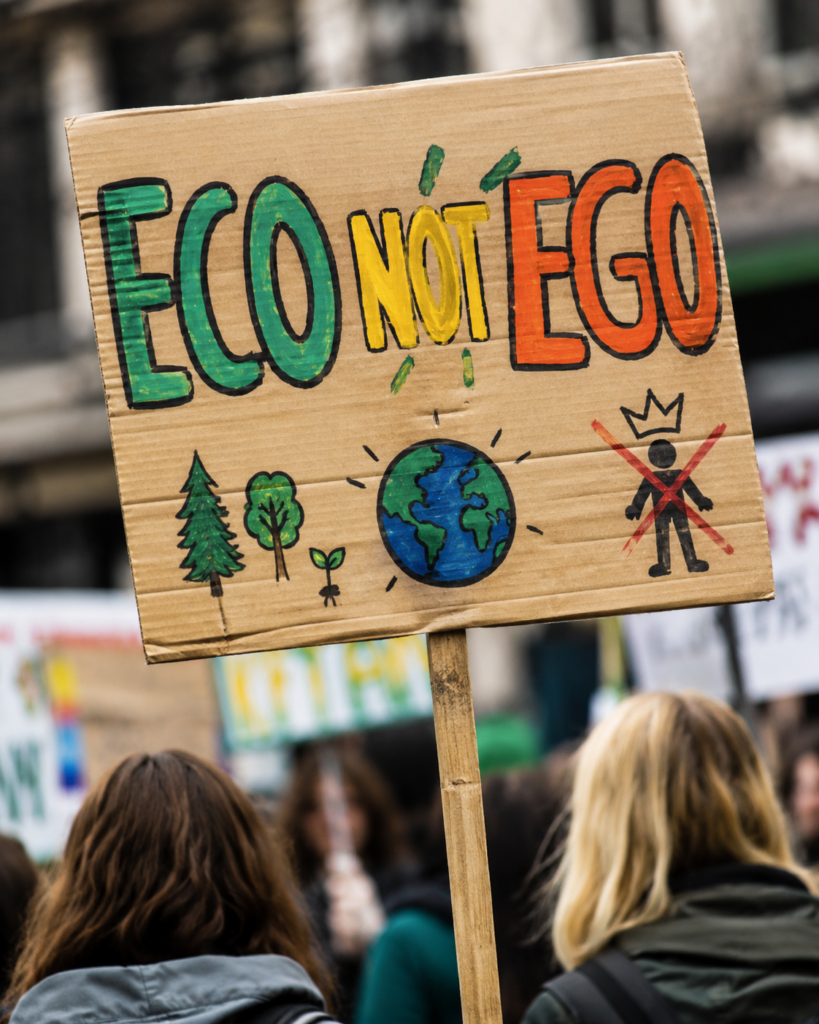 Fotorealistisches Bild einer Klimademonstration. Im Mittelpunkt steht ein selbst gestaltetes Pappschild mit der Aufschrift „ECO NOT EGO“ in grünen, gelben und orangefarbenen Buchstaben. Darunter sind Bäume, eine kleine Pflanze, die Erde sowie eine durchgestrichene Figur mit Krone gezeichnet. Im unscharfen Hintergrund sind weitere Demonstrierende und Protestschilder zu sehen.